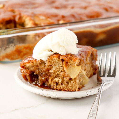 Piece of apple cake on a white plate with a fork topped with a scoop of vanilla ice cream.