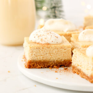 Cheesecake bar topped with whipped cream on a white plate with glass of eggnog in the background.