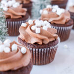 Chocolate cupcake topped with chocolate frosting and mini marshmallows with a Christmas tree in the background.