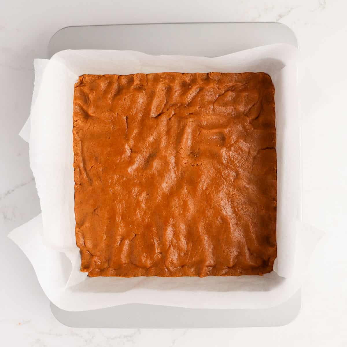 Ginger cookie bar dough pressed into a square metal pan lined with parchment paper.