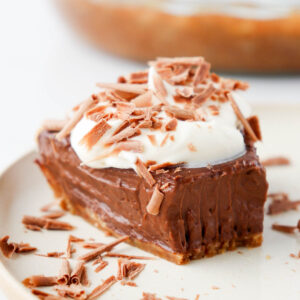 Slice of chocolate pie topped with whipped cream and chocolate shavings on a beige plate.