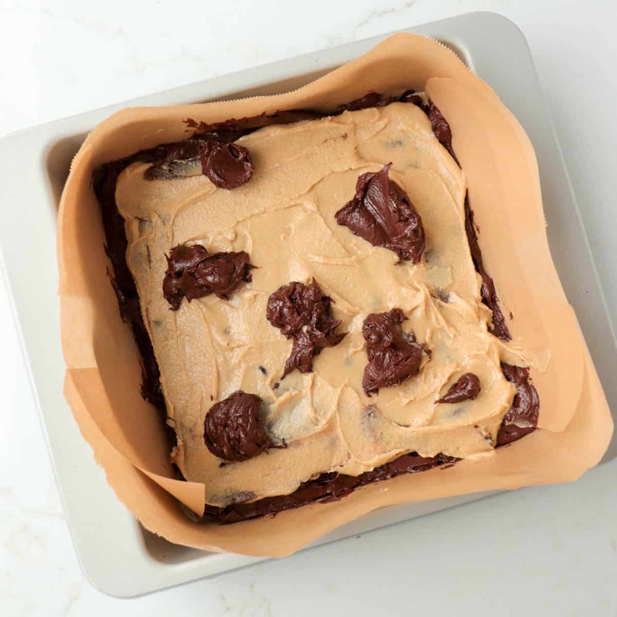 Tahini mixture spread on top of brownie batter in the pan with blobs of remaining batter on top.