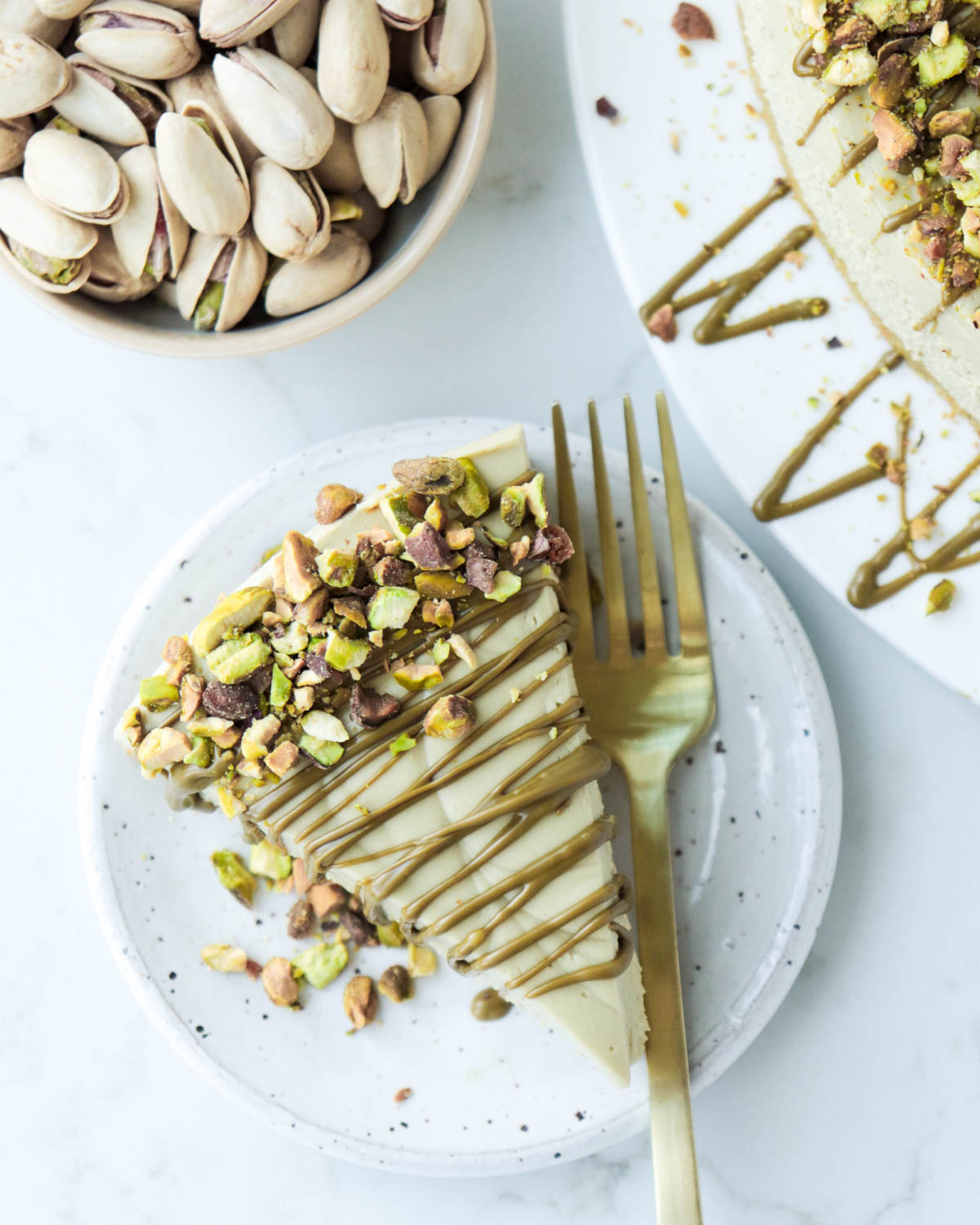 Slice of pistachio cheesecake on a white plate topped with pistachio cream and chopped pistachios.