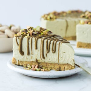 Slice of pistachio cheesecake on a white plate topped with a drizzle of pistachio cream and chopped pistachios.